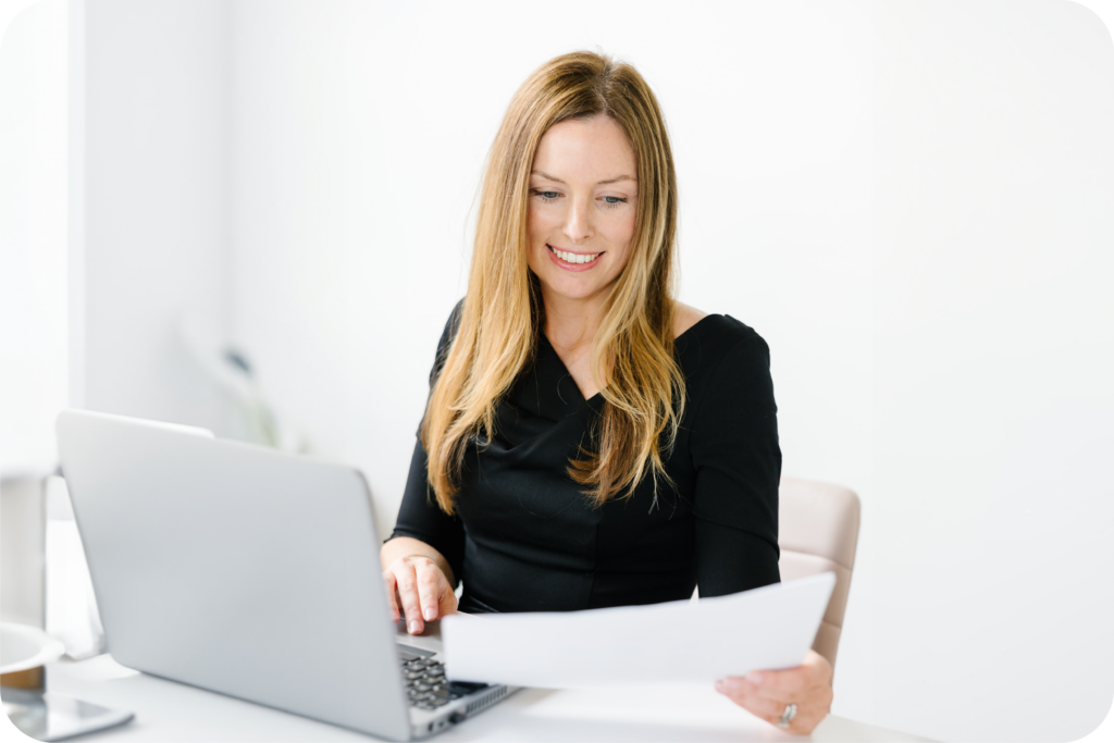 A young woman is a black top smiling, looking over a document while scrolling on her laptop with her other hand.