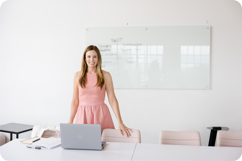 A young woman in a pink dress standing at her desk, while facing the camera. Behind her is a dry erase board with her business SWOT (Strengths, Weaknesses, Opportunities, and Threats) notes.