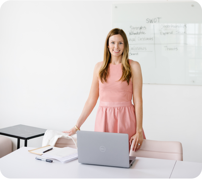 A young woman in a pink dress standing at her desk, while facing the camera. Behind her is a dry erase board with her business SWOT (Strengths, Weaknesses, Opportunities, and Threats) notes.