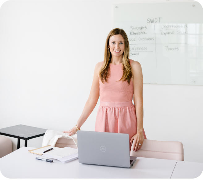 A young woman in a pink dress standing at her desk, while facing the camera. Behind her is a dry erase board with her business SWOT (Strengths, Weaknesses, Opportunities, and Threats) notes.